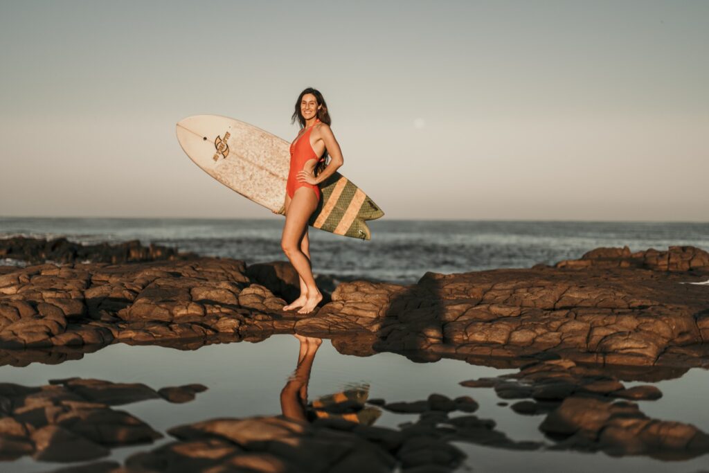 Hannah on the rocks with her surfboard at sunset